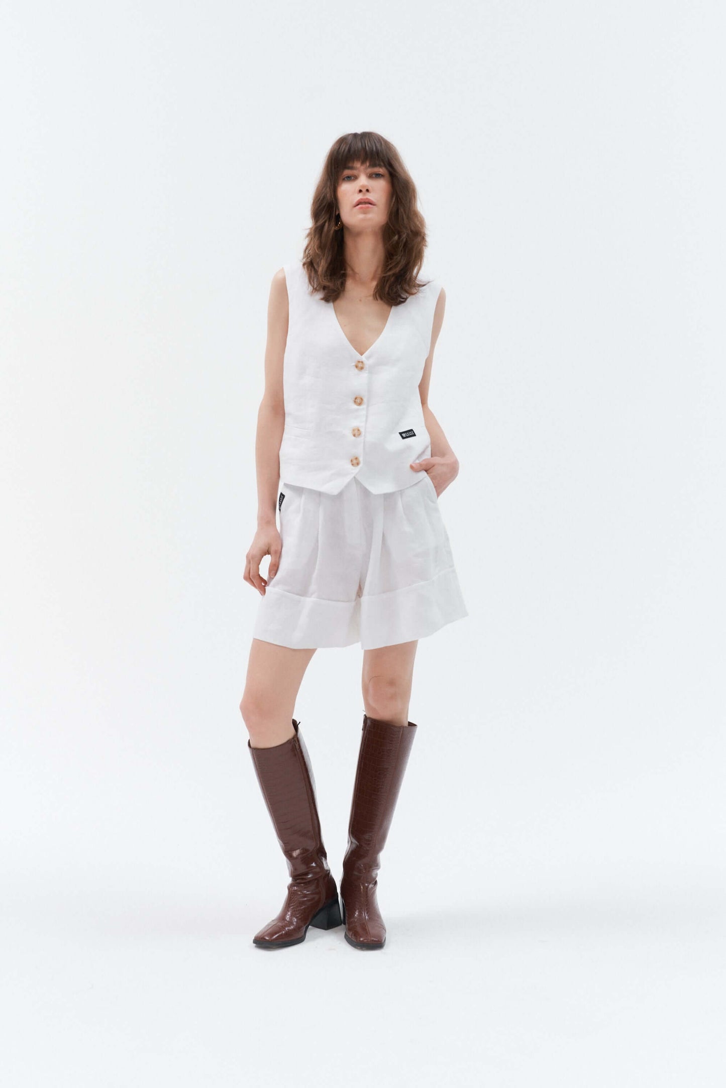 Front view of woman wearing white wide-leg linen shorts Bonita paired with the white linen waistcoat Vesta, styled with the brown high-heeled boots.