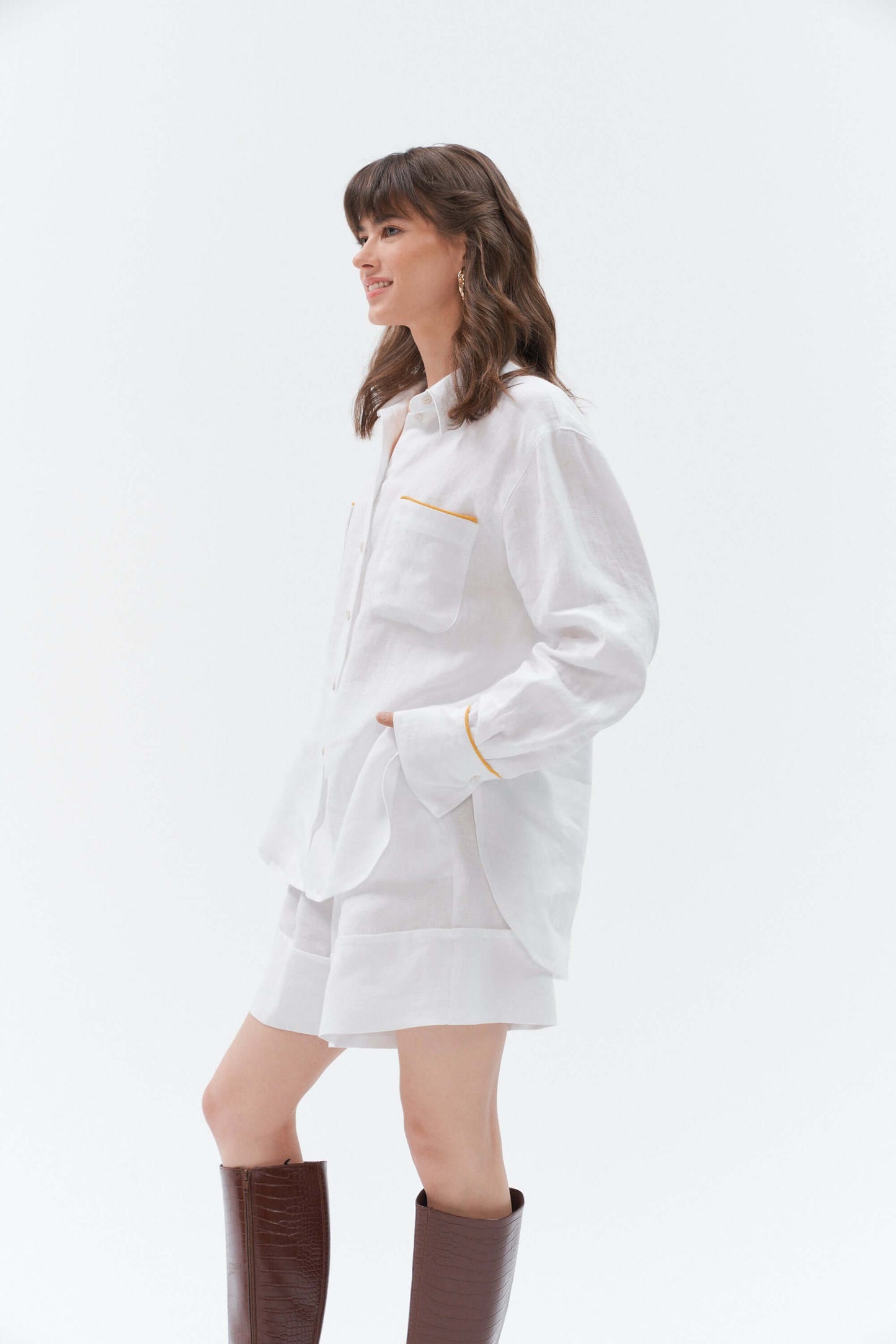 Side view of woman wearing white wide-leg linen shorts Bonita paired with the white Brillante long-sleeve oversized linen shirt with yellow accents, styled with brown high-heeled boots.