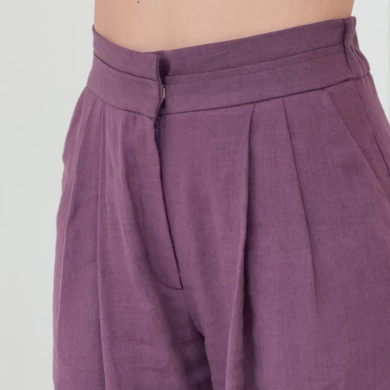 Close-up video of a woman wearing plum-colored high-waisted linen pants, highlighting the elasticated waistband for comfort and the front zipper for a refined, structured look.